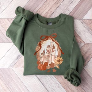 May include: An olive green sweatshirt with a fall-themed illustration. The design features a castle, pumpkin, sunflower, and autumn leaves. The sleeves are cuffed. The sweatshirt is folded on a wooden surface.