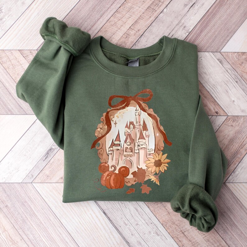 May include: An olive green sweatshirt with a fall-themed illustration. The design features a castle, pumpkin, sunflower, and autumn leaves. The sleeves are cuffed. The sweatshirt is folded on a wooden surface.