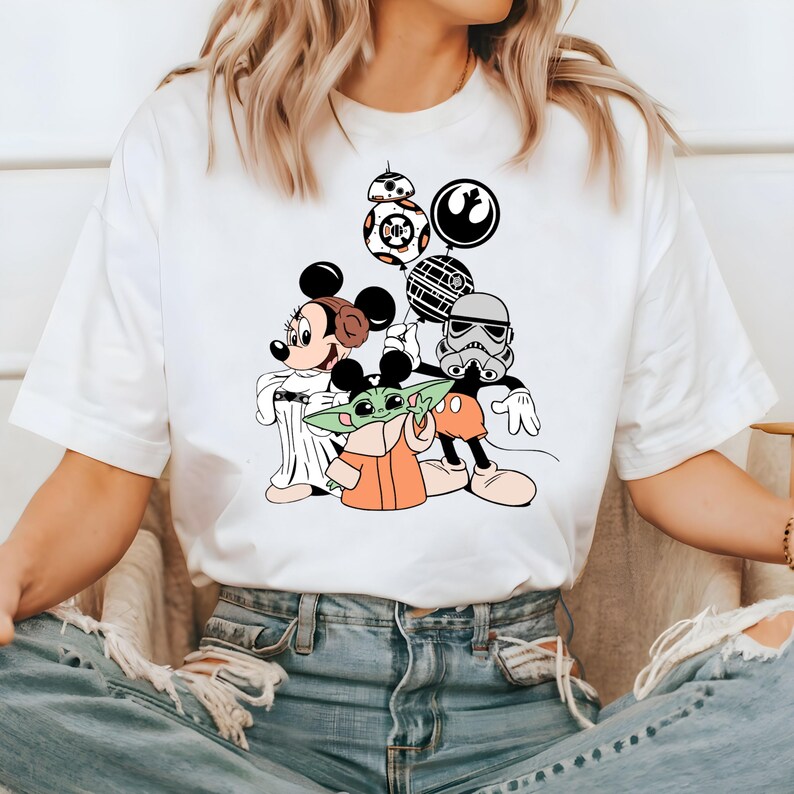 May include: White t-shirt featuring a colorful graphic of Mickey Mouse, Minnie Mouse, Baby Yoda, a Stormtrooper, and BB-8. The design includes Star Wars and Disney elements. The shirt is made of a soft material.