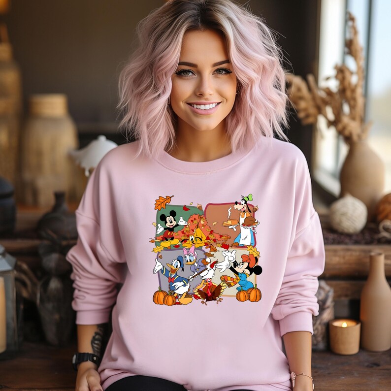 May include: A light pink sweatshirt featuring a colorful graphic of Disney characters celebrating autumn. The graphic includes Mickey Mouse, Minnie Mouse, Donald Duck, and Goofy, surrounded by fall leaves and pumpkins. The sweatshirt is long-sleeved.