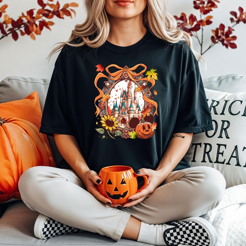 May include: Black t-shirt featuring a colorful graphic of a castle, sunflowers, and a jack-o'-lantern. The person is holding an orange pumpkin-shaped mug. The background includes an orange pillow and a pillow with the text "Trick or Treat".