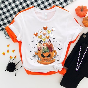 May include: White t-shirt with a graphic of three witches flying in a pumpkin-shaped cup with Mickey Mouse-shaped balloons. The witches are wearing green, purple, and blue dresses. The cup is orange and has a spooky face.