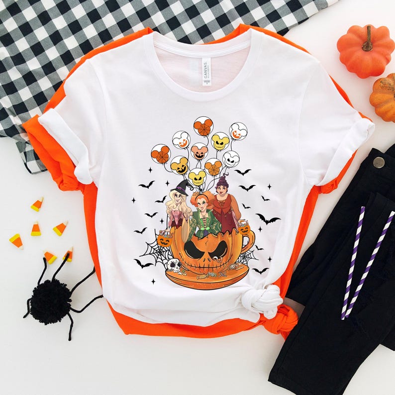 May include: White t-shirt with a graphic of three witches flying in a pumpkin-shaped cup with Mickey Mouse-shaped balloons. The witches are wearing green, purple, and blue dresses. The cup is orange and has a spooky face.
