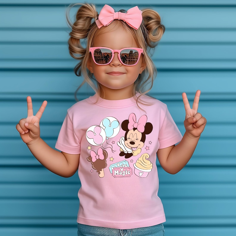 May include: A pink t-shirt with a graphic of Minnie Mouse holding balloons, an ice cream cone, and a ticket that says "Magic".