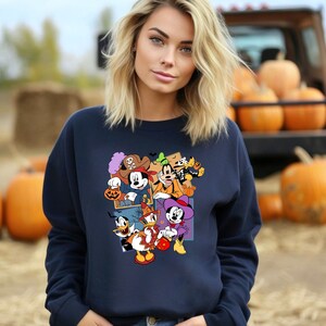 May include: A navy blue sweatshirt with a colorful graphic print of Mickey Mouse and friends dressed in Halloween costumes. The graphic features Mickey Mouse as a pirate, Minnie Mouse as a witch, Donald Duck as a ghost, and Goofy as a skeleton.