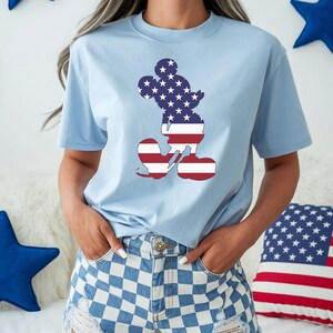May include: Pale blue t-shirt with a Mickey Mouse outline filled with a US flag design. The shirt is worn with blue and white chequered shorts. A red, white, and blue cushion is in the background.