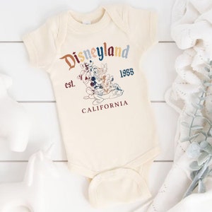 May include: A cream-colored baby onesie with a vintage-style Disneyland logo featuring Mickey Mouse and friends. The text reads "Disneyland est. 1955 CALIFORNIA".