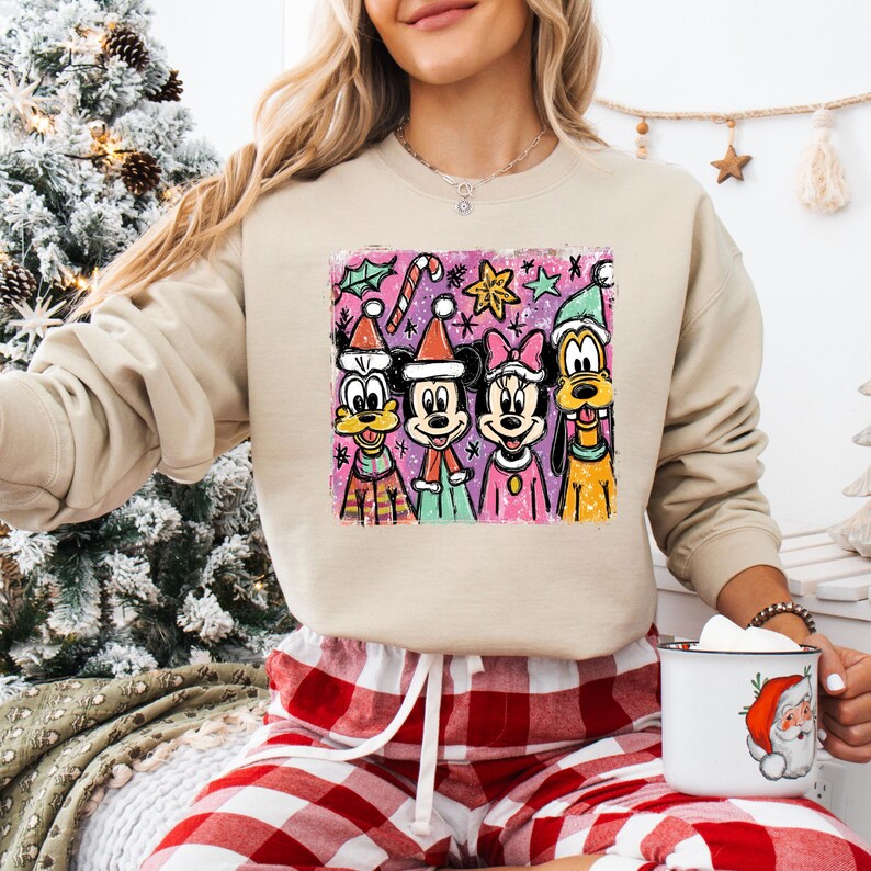 Op de afbeelding: Een beige sweatshirt met een kleurrijke afbeelding van Disney-personages in feestelijke kleding. De afbeelding bevat Mickey Mouse, Minnie Mouse, Goofy en Donald Duck, met feestelijke elementen zoals zuurstokken en sterren.