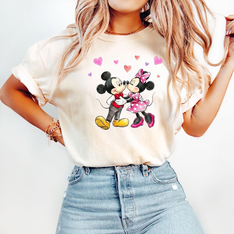May include: A cream-coloured t-shirt with a watercolour illustration of Mickey and Minnie Mouse embracing. Minnie wears a pink polka-dot dress and bow. Hearts float above the couple. The shirt is a casual style.