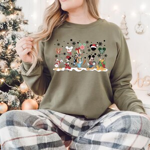 May include: A green sweatshirt with a Christmas-themed design featuring Mickey Mouse and his friends. The design includes Mickey Mouse, Minnie Mouse, Donald Duck, Goofy, Pluto, and a snowman. The characters are holding balloons and gifts. The design is surrounded by snowflakes.