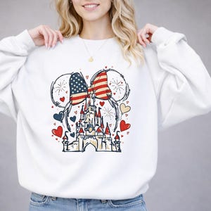 May include: White sweatshirt featuring a graphic of Mickey Mouse ears with a bow in the American flag design. A castle is centered below the ears, surrounded by red, blue, and yellow hearts. Fireworks are also included in the design.