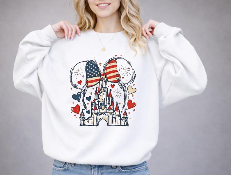 May include: White sweatshirt featuring a graphic of Mickey Mouse ears with a bow in the American flag design. A castle is centered below the ears, surrounded by red, blue, and yellow hearts. Fireworks are also included in the design.