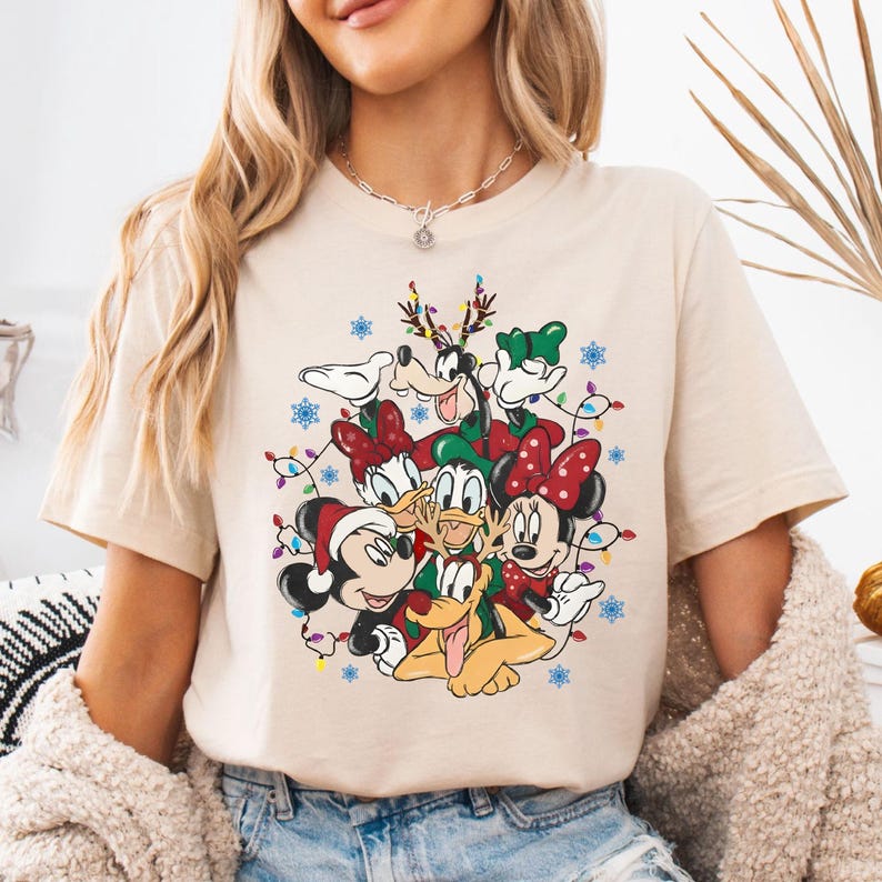 May include: Beige t-shirt featuring a festive graphic of Disney characters, including Mickey and Minnie Mouse, Donald Duck, and Goofy, adorned with Christmas lights and snowflakes. The characters are wearing Santa hats and reindeer antlers.