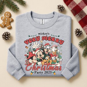 Puede incluir: Sudadera gris claro con un dise&ntilde;o festivo de Mickey Mouse y sus amigos con atuendos navide&ntilde;os. El dise&ntilde;o incluye el texto "mickey's VERY MERRY Christmas Party 2025" en rojo y verde. Prenda de tem&aacute;tica navide&ntilde;a.