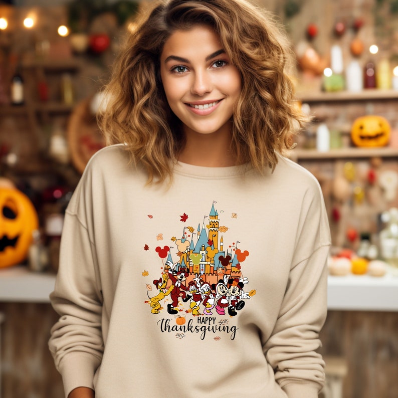 May include: A beige sweatshirt with a graphic print of Mickey Mouse and friends in front of a cartoon version of the Cinderella Castle. The graphic also includes the text "Happy Thanksgiving".