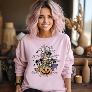 May include: A light pink sweatshirt featuring a Halloween-themed graphic of Disney characters including Mickey Mouse, Minnie Mouse, Donald Duck, and Goofy. The design includes a carved pumpkin and bats.