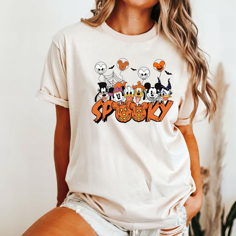 May include: Cream-coloured t-shirt with a Halloween graphic. The design features cartoon characters, jack-o'-lanterns, and the word "Spooky" in orange. The characters are dressed in Halloween costumes, with bats and spiderwebs.