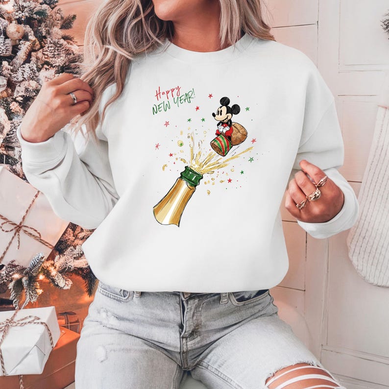 May include: White sweatshirt with a festive design featuring a champagne bottle and Mickey Mouse. The words "Happy New Year!" are written in green. The design includes red and green stars, creating a celebratory theme.