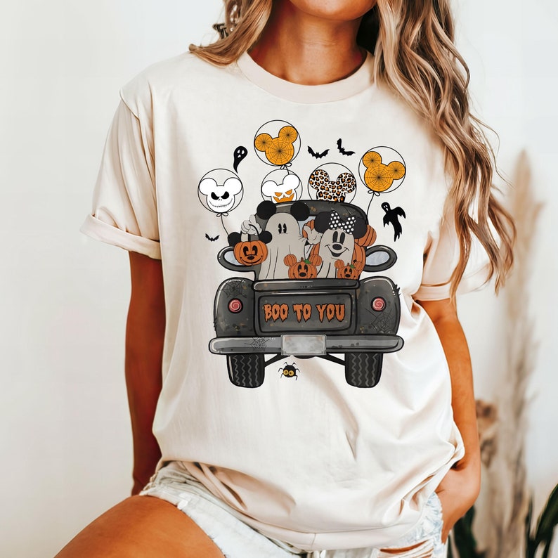 Peut inclure: Un t-shirt blanc avec une illustration noire et blanche de Mickey Mouse, Minnie Mouse et un fant&ocirc;me dans un pick-up noir avec des citrouilles. Le camion porte l'inscription "BOO TO YOU" &agrave; l'arri&egrave;re. Les personnages tiennent des ballons avec des oreilles de Mickey Mouse.