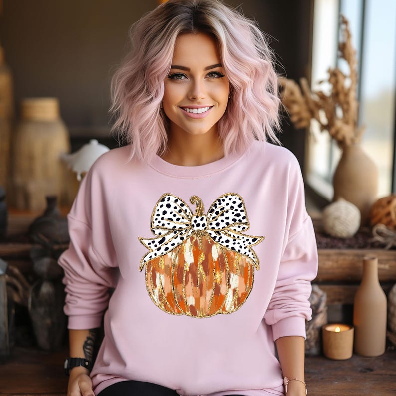 Puede incluir: Una sudadera rosa con un gr&aacute;fico de una calabaza con un lazo de lunares blanco y negro. La calabaza es naranja y marr&oacute;n con detalles de purpurina dorada.
