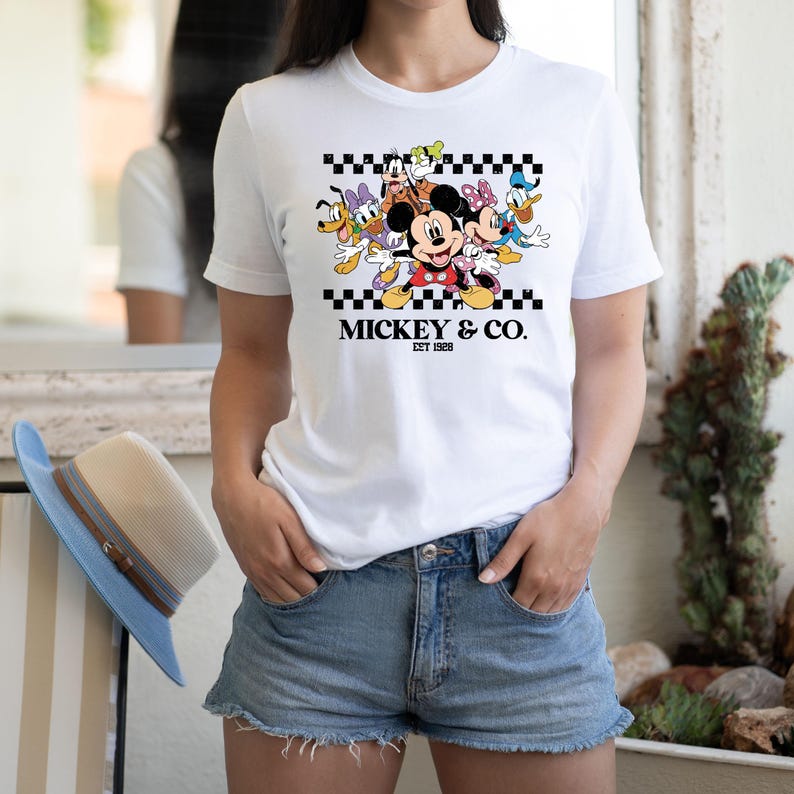 May include: White t-shirt featuring a graphic of Mickey Mouse and friends, with the text "Mickey & Co. EST 1928" in black. The design includes a black and white checkered pattern. The shirt is paired with denim shorts.