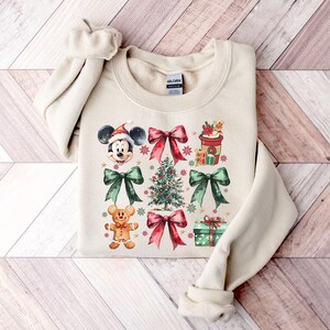 May include: A beige sweatshirt with a Christmas-themed design featuring Mickey Mouse, a gingerbread man, a Christmas tree, bows, and a coffee cup. The design is in watercolor style with red, green, and gold accents.
