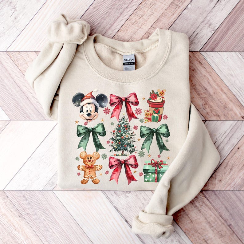 May include: A beige sweatshirt with a Christmas-themed design featuring Mickey Mouse, a gingerbread man, a Christmas tree, bows, and a coffee cup. The design is in watercolor style with red, green, and gold accents.
