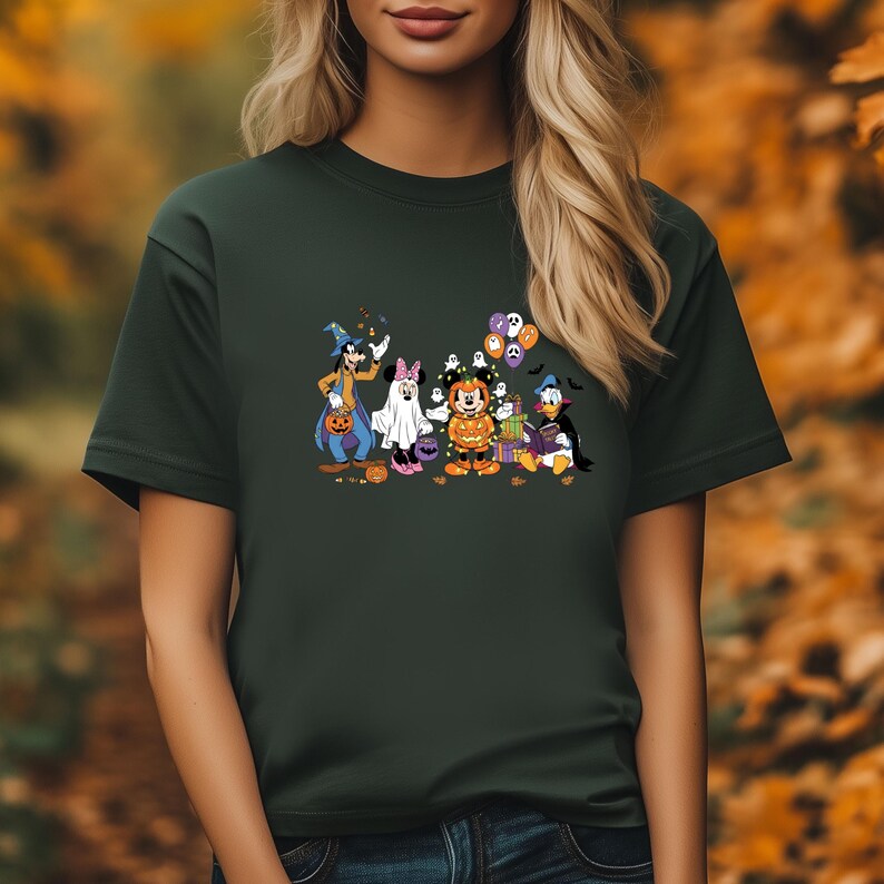 May include: Dark green t-shirt featuring a Halloween-themed graphic. The graphic includes cartoon characters dressed in costumes, such as a witch, a ghost, and a pumpkin. The characters are in front of a blurred autumn background.