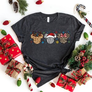 May include: A dark gray t-shirt with a Christmas-themed design featuring three cartoon animal heads. The heads are a reindeer with antlers and lights, a zebra wearing a Santa hat, and a leopard with a red bow. The design is centered on the shirt.