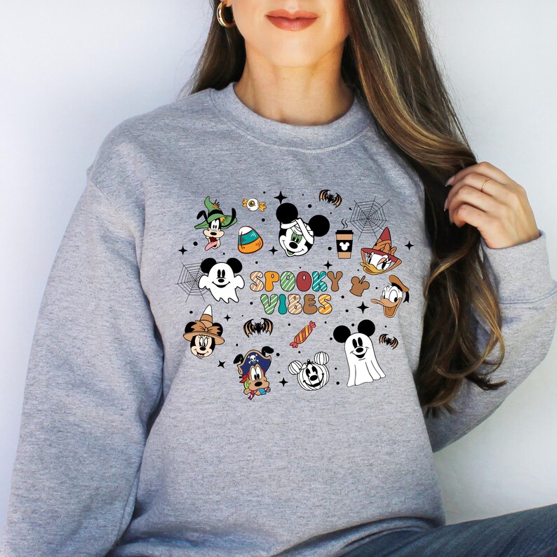 May include: A grey sweatshirt featuring a colourful graphic print of various Disney characters dressed in Halloween costumes. The text "Spooky Vibes" is printed in colourful letters on the sweatshirt.