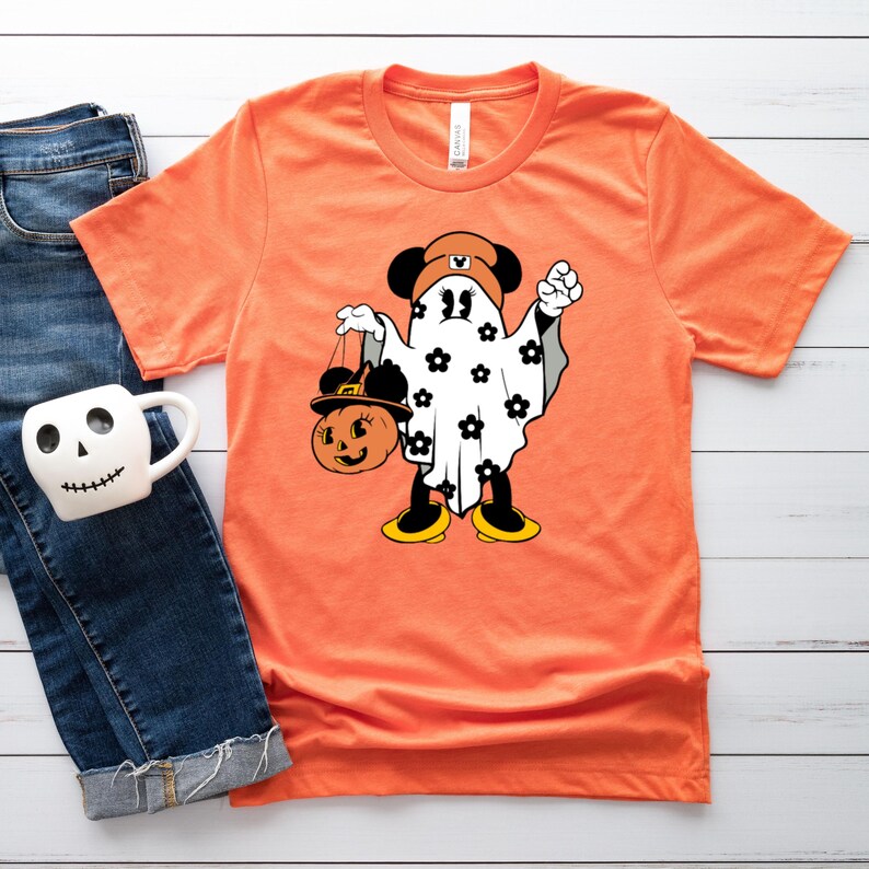 May include: Orange t-shirt featuring a cartoon ghost character with black ears, a brown hat, and a pumpkin-shaped treat bag. The ghost is decorated with black flower accents. The shirt is displayed on a white wooden surface with a pair of blue jeans and a skull-shaped mug.
