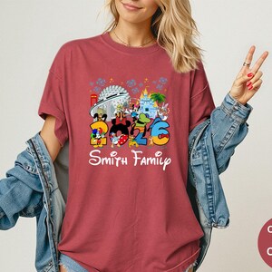 May include: Crimson-colored t-shirt featuring a colorful graphic with the text "2026 Smith Family" and Disney characters. The design includes iconic park landmarks. The shirt is a Comfort Colors brand.