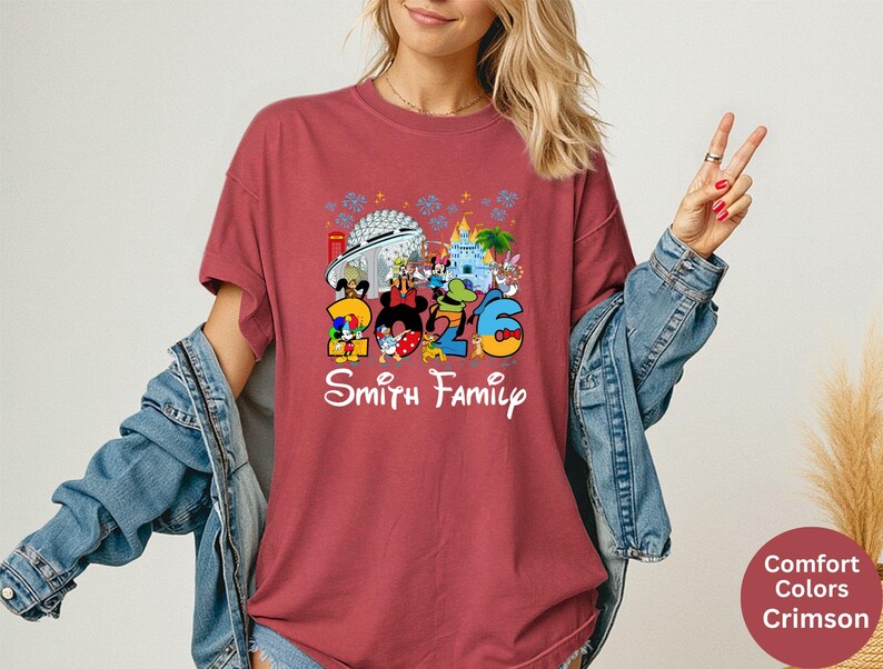 May include: Crimson-colored t-shirt featuring a colorful graphic with the text "2026 Smith Family" and Disney characters. The design includes iconic park landmarks. The shirt is a Comfort Colors brand.