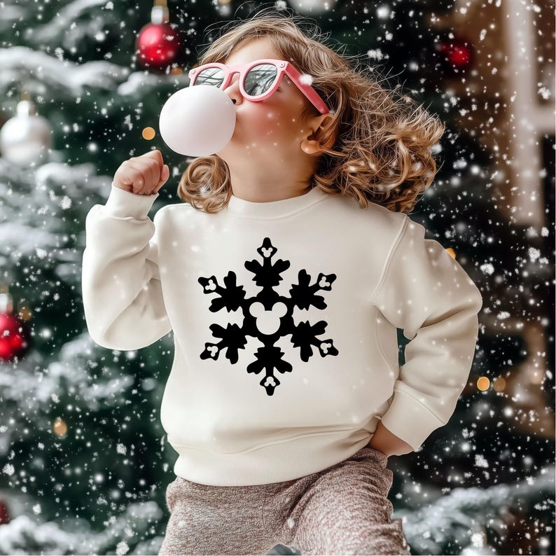 May include: A cream-colored sweatshirt featuring a black snowflake design with a Mickey Mouse head in the center. The sweatshirt is worn by a child wearing pink sunglasses and blowing a bubble. The background includes a Christmas tree and falling snow.