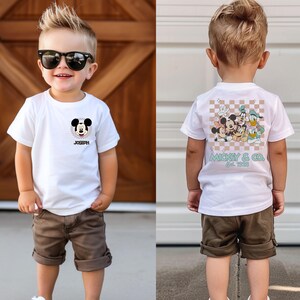 May include: White t-shirt with a Mickey Mouse graphic on the front and a Mickey Mouse and friends graphic on the back. The shirt says "Mickey & Co. Est. 1928" on the back. The shirt is being worn by a young child.