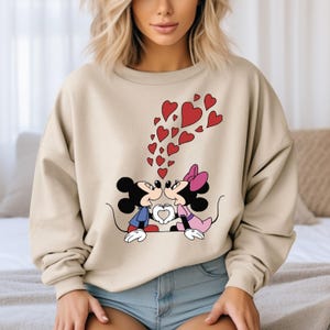 May include: A beige sweatshirt with a graphic print of Mickey Mouse and Minnie Mouse kissing. Red hearts are floating above them.