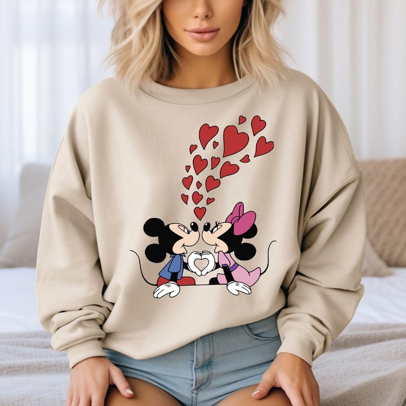 May include: A beige sweatshirt with a graphic print of Mickey Mouse and Minnie Mouse kissing. Red hearts are floating above them.
