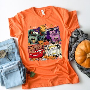 May include: Orange t-shirt with a Halloween-themed graphic featuring cartoon cars dressed up in costumes. The cars are Lightning McQueen, Mater, and a few others. The graphic also includes pumpkins, bats, and other Halloween decorations.