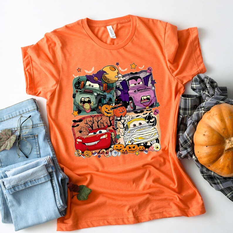 May include: Orange t-shirt with a Halloween-themed graphic featuring cartoon cars dressed up in costumes. The cars are Lightning McQueen, Mater, and a few others. The graphic also includes pumpkins, bats, and other Halloween decorations.