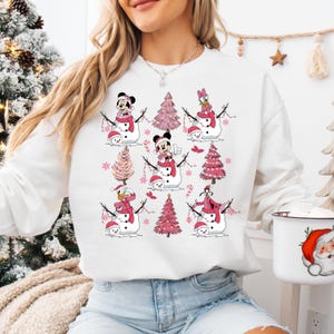 May include: White sweatshirt with a festive design featuring cartoon characters as snowmen. Each snowman has a red scarf and hat, with pink Christmas trees and snowflakes. Characters include Mickey and Minnie Mouse, Donald Duck, and Goofy.