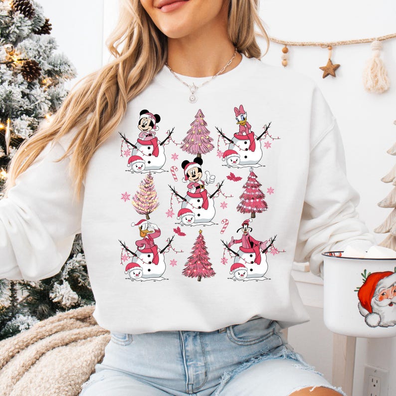 May include: White sweatshirt with a festive design featuring cartoon characters as snowmen. Each snowman has a red scarf and hat, with pink Christmas trees and snowflakes. Characters include Mickey and Minnie Mouse, Donald Duck, and Goofy.