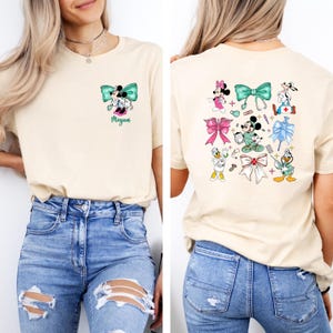 May include: Cream-colored t-shirt featuring a front and back design. The front displays a Minnie Mouse and Mickey Mouse graphic with a teal bow and the name "Megan". The back showcases various cartoon characters with bows and medical-themed elements.