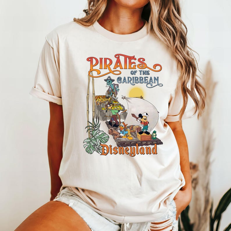 May include: A white t-shirt with a vintage-style graphic of the Pirates of the Caribbean ride at Disneyland. The graphic features Mickey Mouse and other Disney characters on a pirate ship.