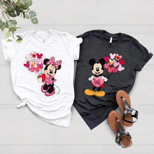 May include: Two white and black t-shirts with a Mickey Mouse and Minnie Mouse design. Mickey Mouse is holding a heart-shaped box of chocolates and Minnie Mouse is holding a bunch of heart-shaped balloons.