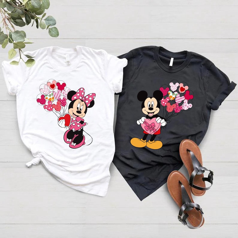 May include: Two white and black t-shirts with a Mickey Mouse and Minnie Mouse design. Mickey Mouse is holding a heart-shaped box of chocolates and Minnie Mouse is holding a bunch of heart-shaped balloons.