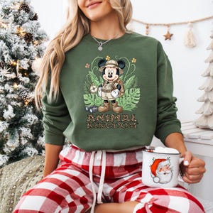 May include: Green sweatshirt featuring a Mickey Mouse safari design with the text "ANIMAL KINGDOM". The graphic includes Mickey in a safari outfit, surrounded by tropical leaves and flowers. The person is wearing red and white plaid pajama pants.