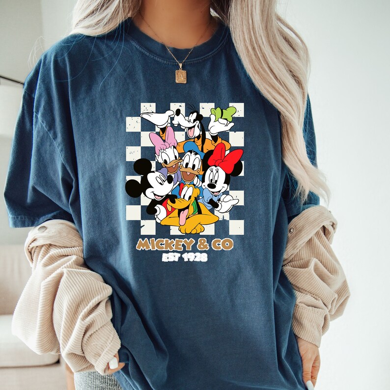 Puede incluir: Una camiseta azul marino con un fondo a cuadros con Mickey Mouse y sus amigos. El texto "Mickey & Co. Est 1928" est&aacute; impreso debajo de la imagen.