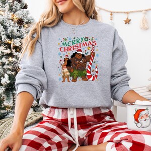 May include: A light gray sweatshirt featuring a festive "Merry Christmas" design with cartoon characters. The graphic includes a candy cane, stars, and the characters wearing Santa hats. The sweatshirt is paired with red and white plaid pajama pants.