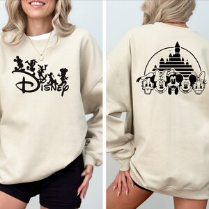 Puede incluir: Una sudadera beige con un logotipo de Disney negro y personajes en la parte delantera y una silueta negra del castillo de Disney y personajes en la parte posterior.