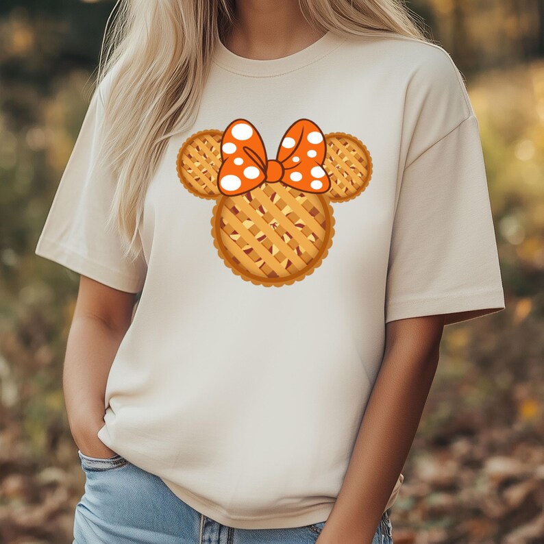 K&ouml;nnte beinhalten: Beiges T-Shirt mit einem Design zum Thema Kuchen. Das Design umfasst einen kuchensf&ouml;rmigen Kopf mit Gitterkrustenohren und einer Schleife mit wei&szlig;en Tupfen. Das T-Shirt hat eine neutrale Farbe und kurze &Auml;rmel.